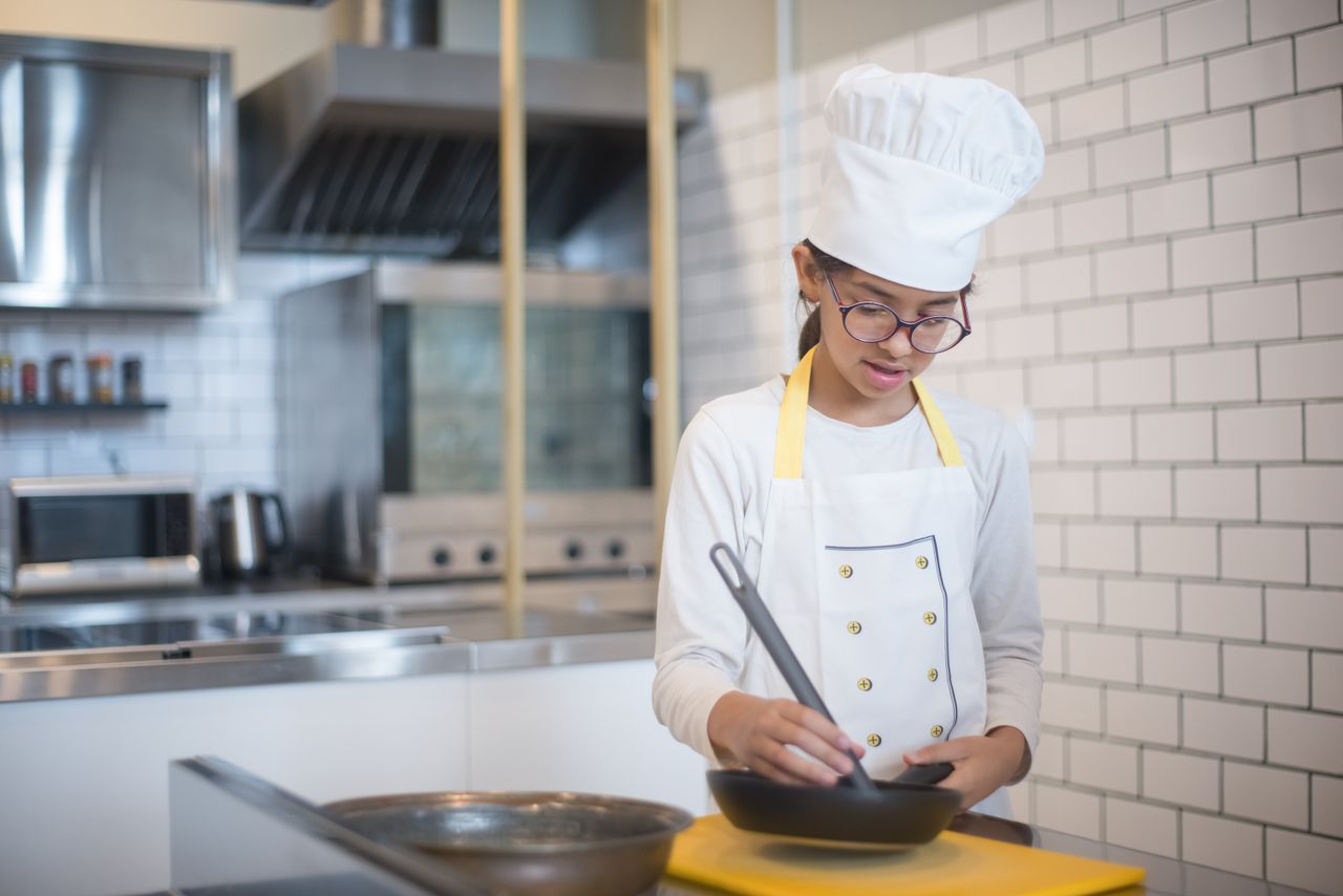 A young girl wearing chef attire cooking in a modern kitchen with a pan.
